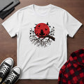 White t-shirt with a red sun and black tree design on a wooden surface with shoes and a plaid shirt.